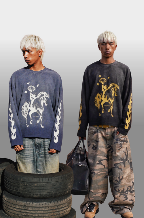 Two male models with platinum blonde hair pose in front of a light gray gradient background, styled with tires and a black duffel bag. The model on the left wears a faded navy blue sweatshirt with a white horse graphic and white flame details, paired with faded blue jeans.