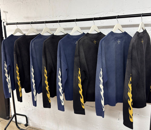 A rack of long-sleeve crewneck sweatshirts in navy, black, and blue, featuring a white wave graphic on the sleeve and a yellow flame motif on the lower sleeve, displayed against a white brick wall.